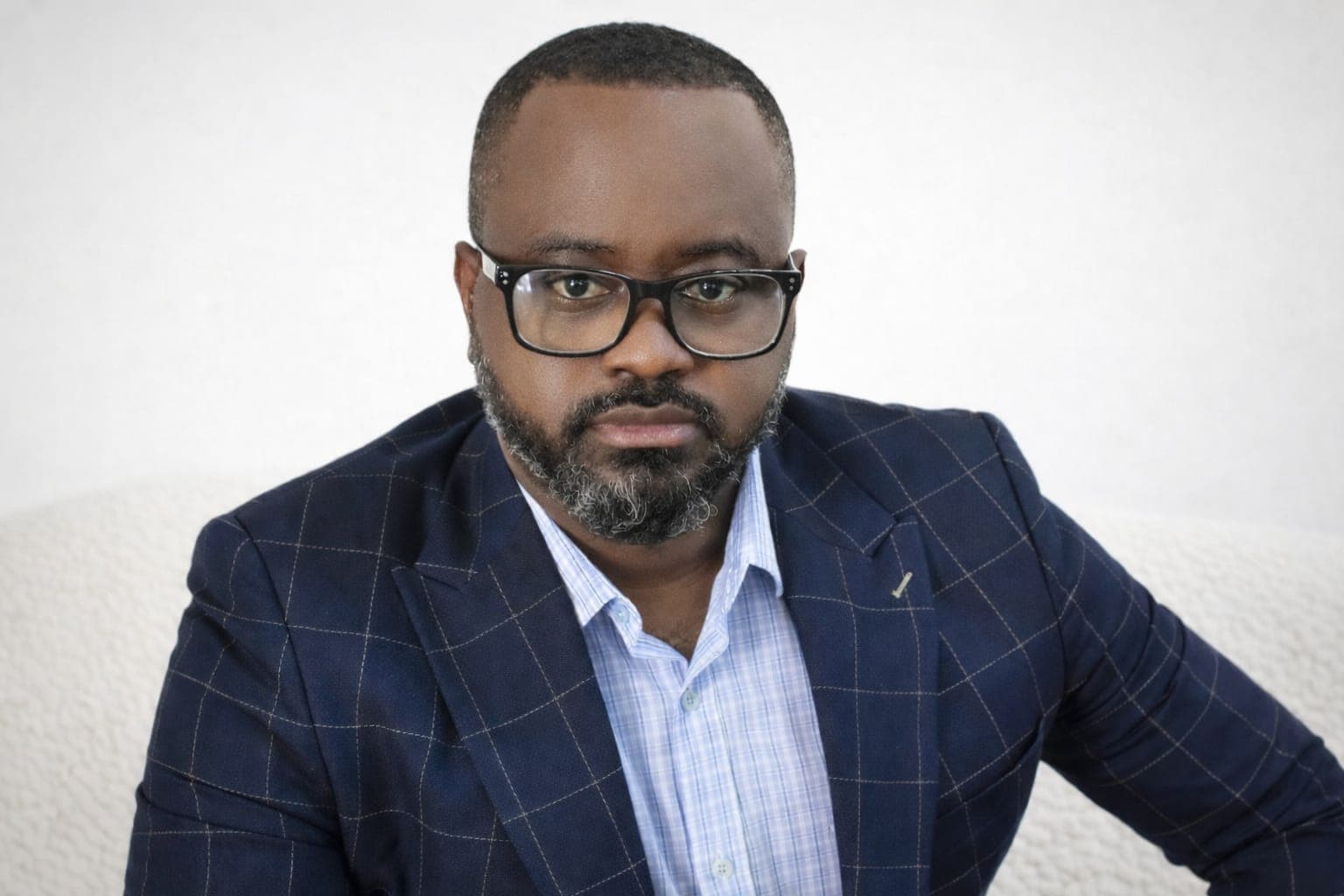 Black man with glasses and a beard wearing a blue windowpane blazer and light shirt.